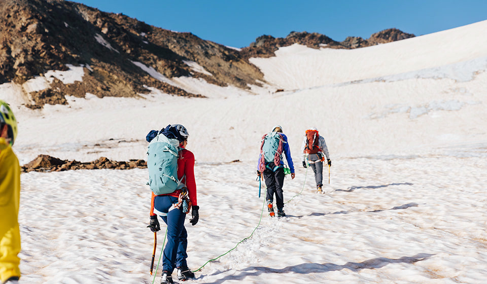 Three female mountaineers with backpacks and crampons walk across a snowfield towards a mountain peak.
