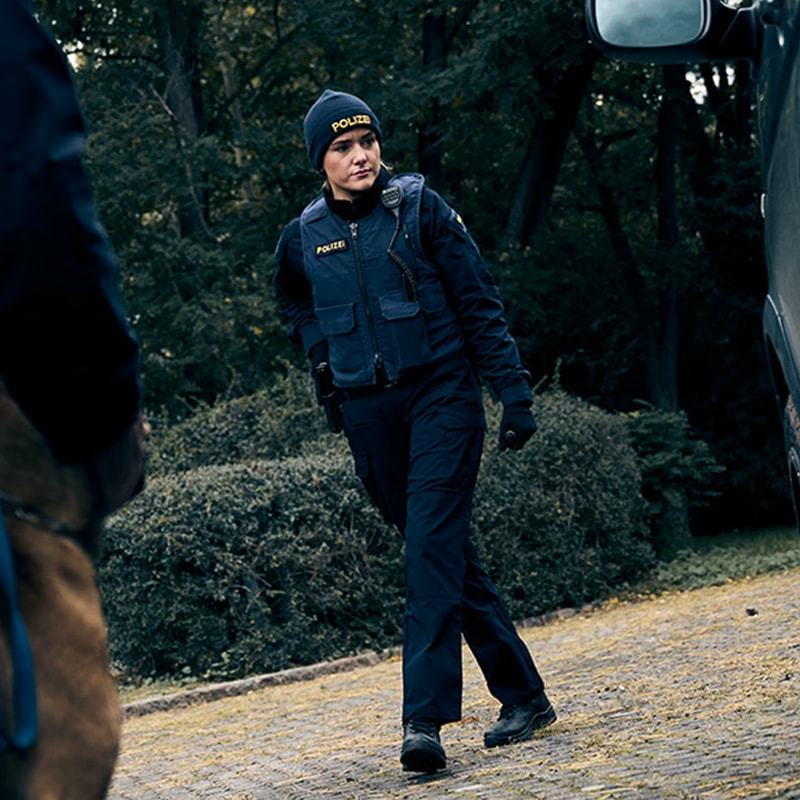 Deux policiers, dont l’un avec un chien d’assistance, s’approchent d’une femme en uniforme policier. Elle se tient sur un chemin pavé à côté d’un véhicule sombre. L’arrière-plan est une forêt sombre. La scène semble mystérieuse et sérieuse.