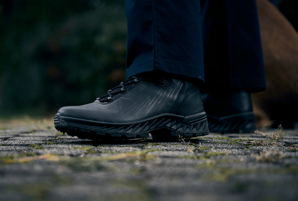 Chaussure noire pour homme robuste avec laçage et semelle grossière, posée sur un chemin pavé.