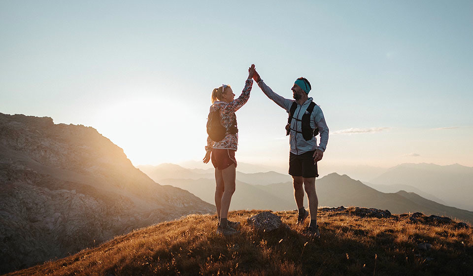 Zwei LOWA Trailrunner auf einem Berggipfel bei Sonnenuntergang, die sich ein High-Five geben.