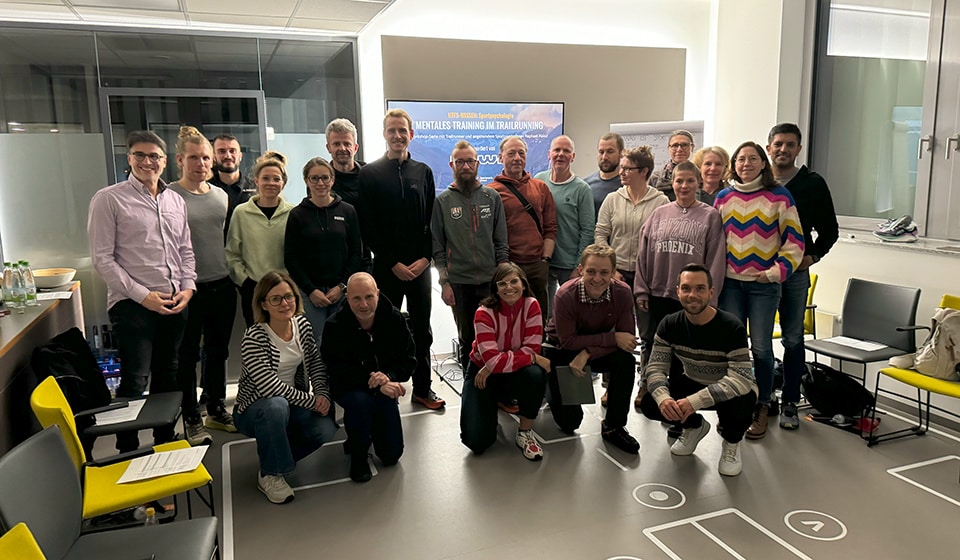 Un gruppo di 16 persone si alza e si siede in una sala di allenamento, uno schermo sullo sfondo mostra la scritta tedesca.