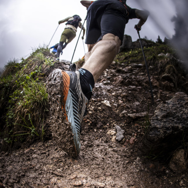 Runner on muddy mountain trail, focus on dirty shoe and hiking poles.