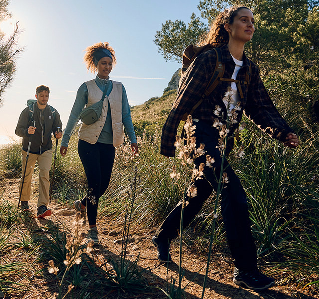 Three hikers in light clothing walk on a sunny dirt road.