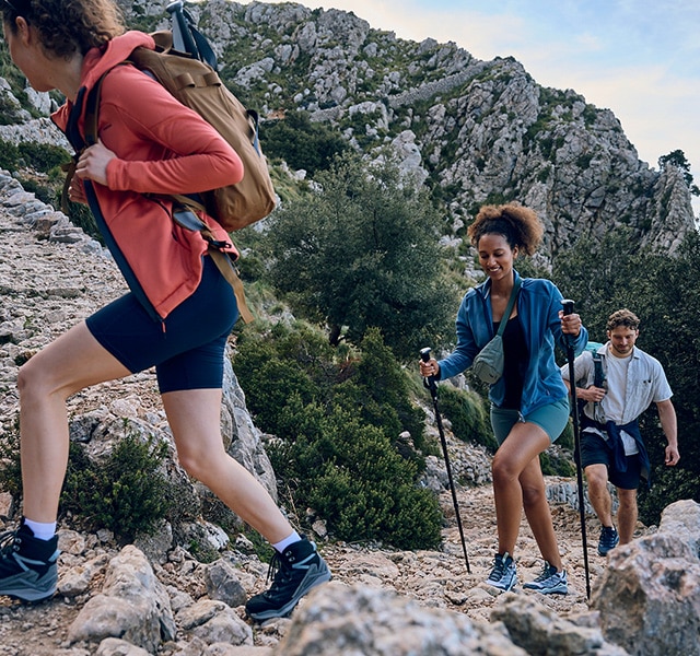 Three hikers climb up a rocky mountain path. A woman in red in front, a woman in blue in the middle, a man in white in the back.