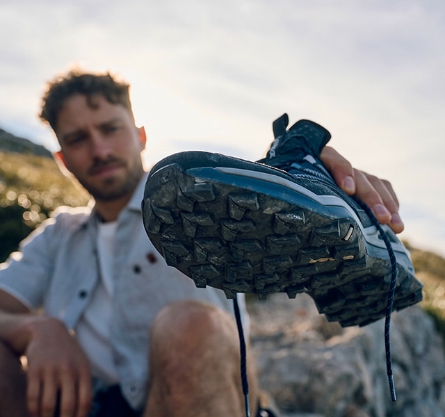 A man holds a muddy hiking boot up to the camera, his face blurred in the background.