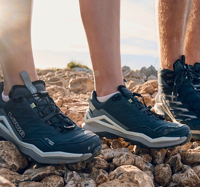 Feet in LOWA hiking boots on stony ground, sunset background