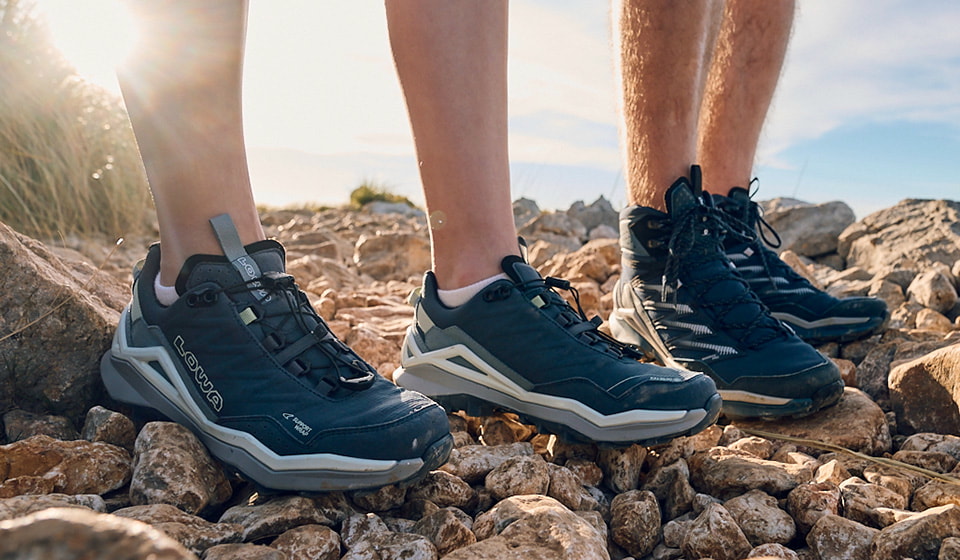 Man holds a dirty hiking boot with a coarse sole in the camera, mountains and sky in the background.