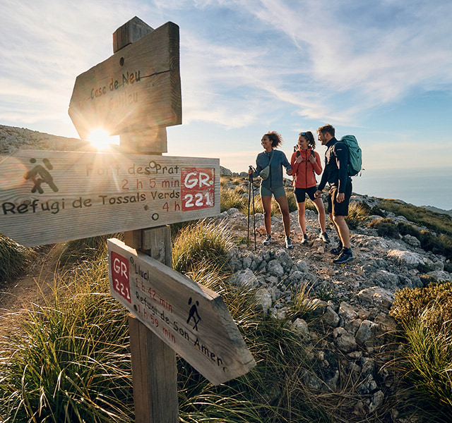 Hiking trail sign with three hikers in front of a mountainous backdrop at sunset.