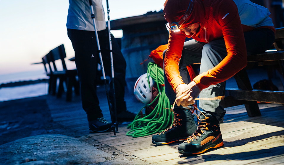 Close-up of hiking boots in shades of blue on rocky ground. In the background, a person in a yellow jacket is walking.