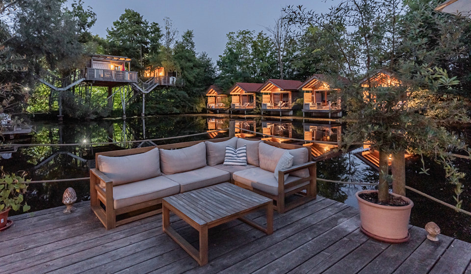Eine Lounge-Ecke auf einer Terrasse vor einem kleinen See. Im Hintergrund sind kleine, hölzerne Ferienhäuser zu sehen.