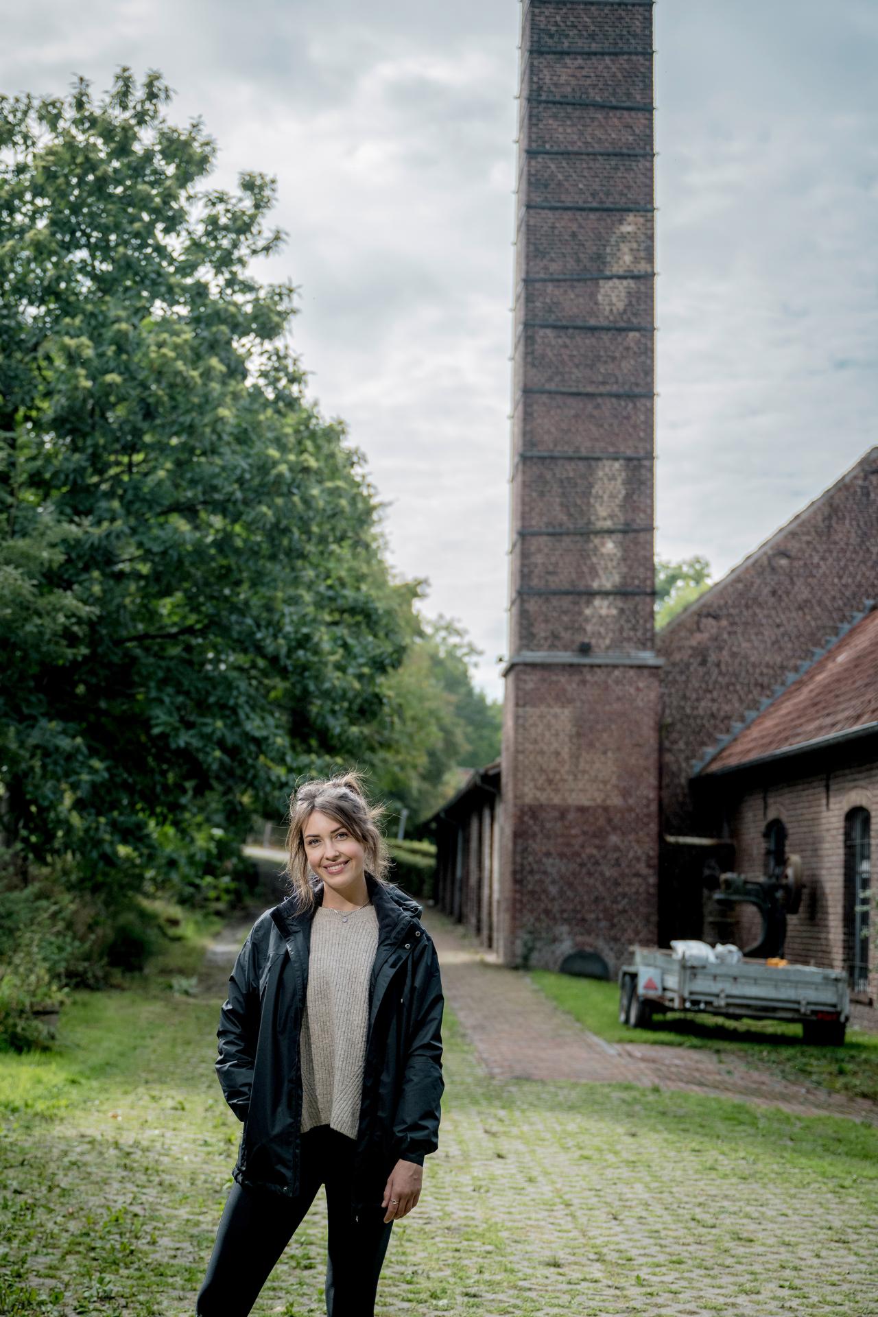 Eine lächelnde Frau steht vor einem Backsteinturm und einem Gebäude mit Ziegeldach.