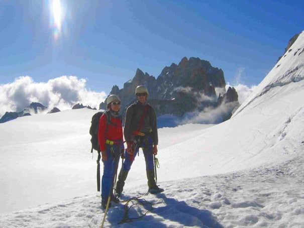 Two mountaineers in full gear stand on a snow-covered peak, rugged rock formations and cloudy skies in the background.