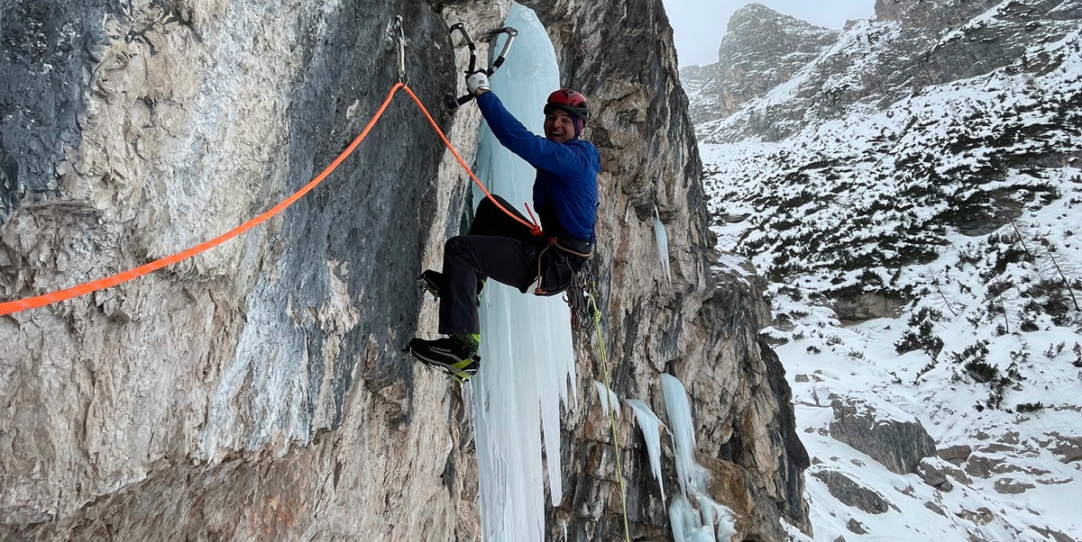 An ice climber with a helmet and ice tools hangs from a rock face, next to icy stalactites.