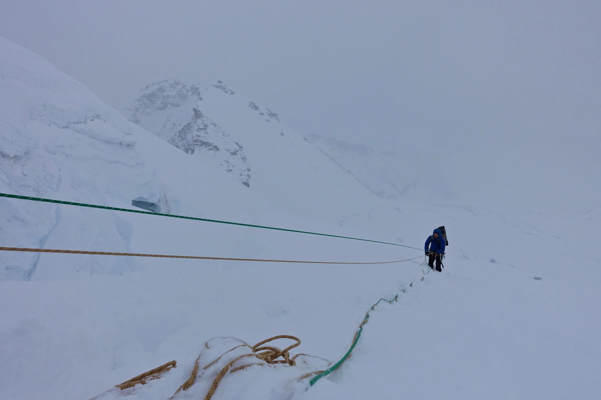 Bergsteiger in blauer Kleidung steigt auf verschneiter Bergflanke an Seilen entlang, Nebel.