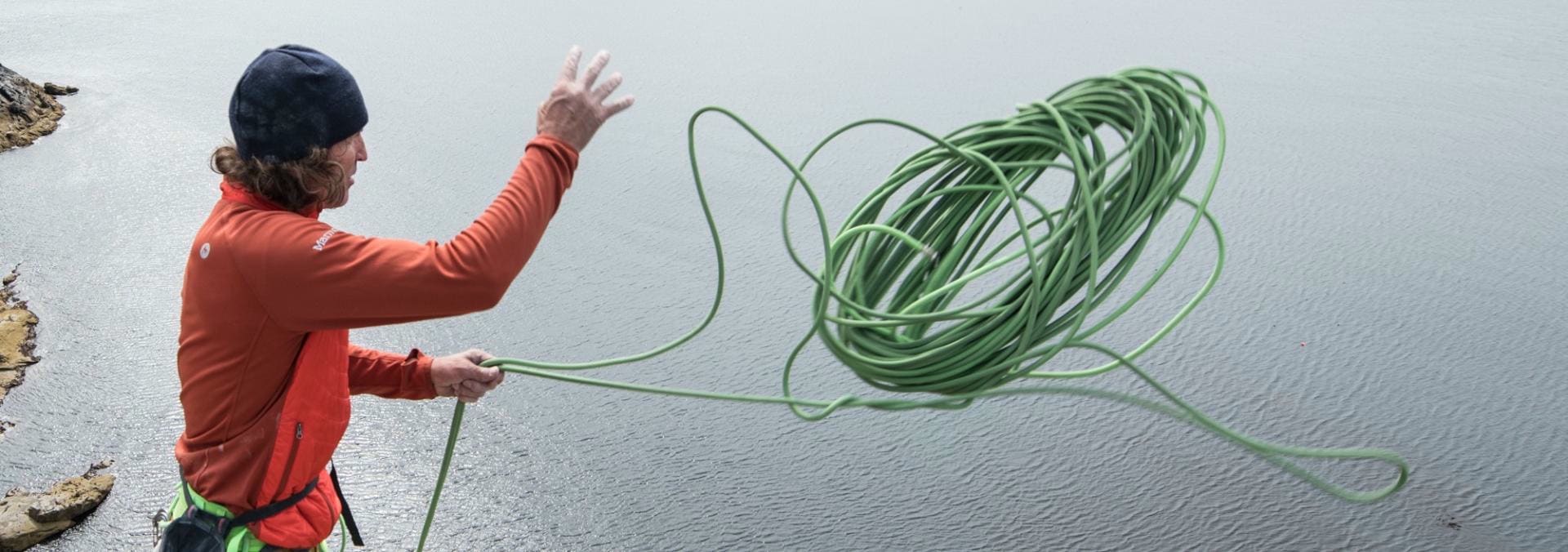 A mountaineer in a red sweater and black cap throws a green rope over calm water.