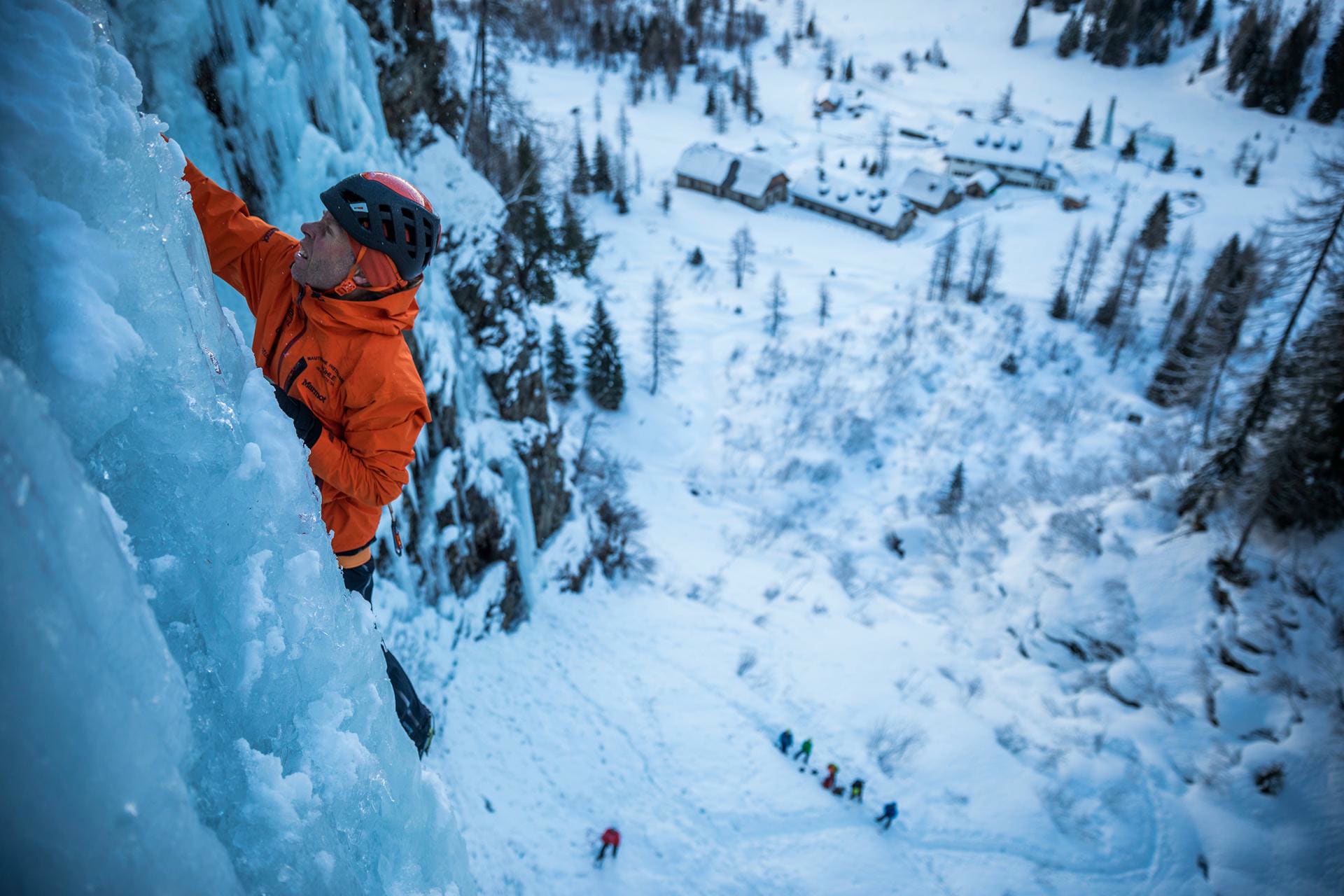 Icy climb: Mountaineer in orange jacket climbs on a frozen waterfall, building and hikers in the snow below.