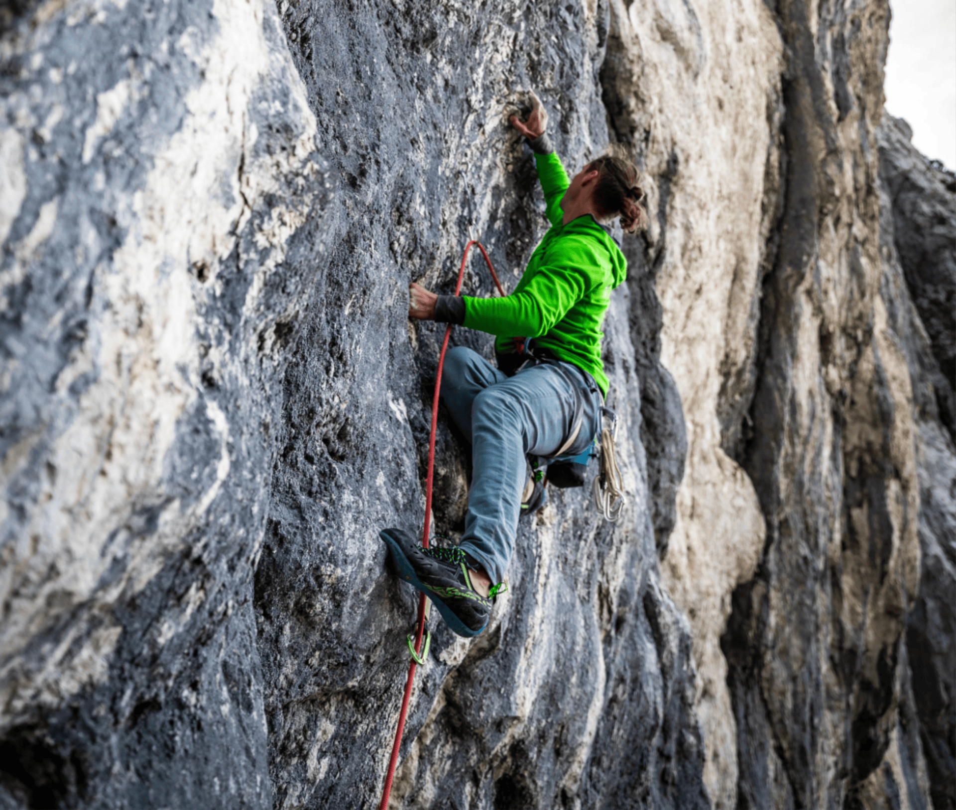 Ein Kletterer in grüner Jacke klettert an einer grauen Felswand, gesichert durch ein rotes Seil.