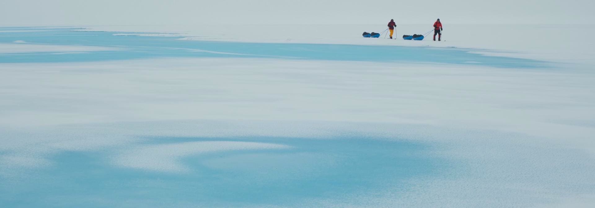 Two cross-country skiers with sleds on ice surface under bright sky.