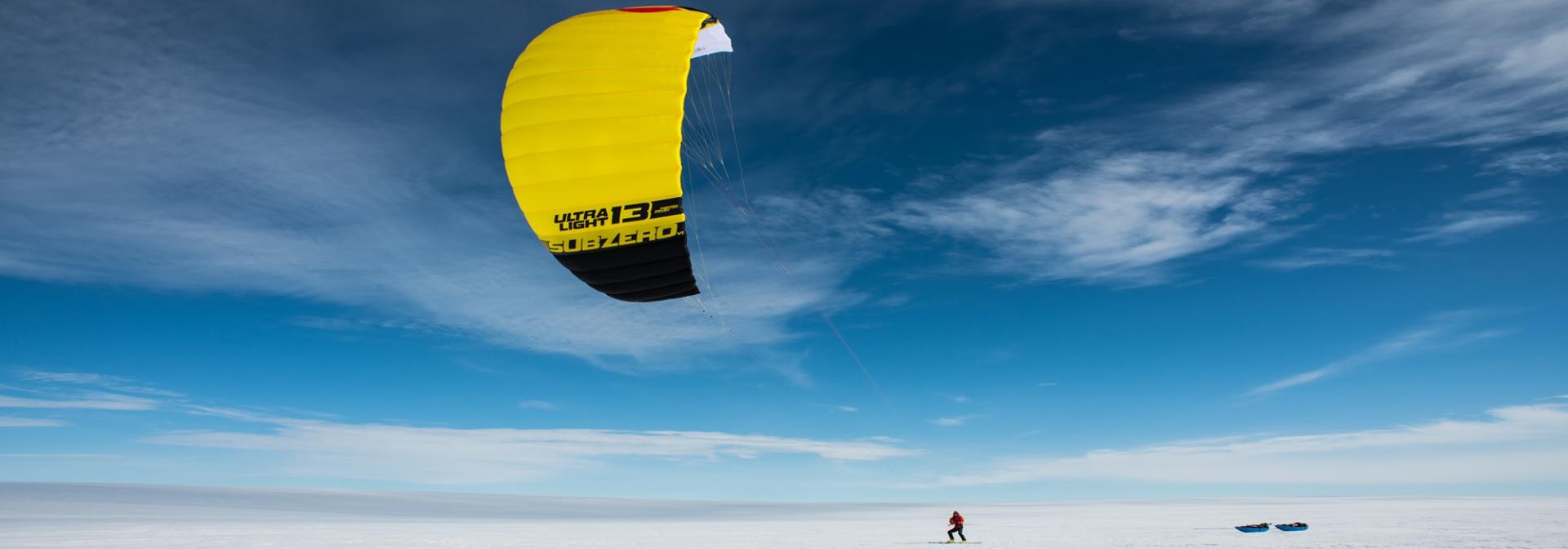 A man on skis under a yellow kite in an icy landscape with blue skies and thin clouds.