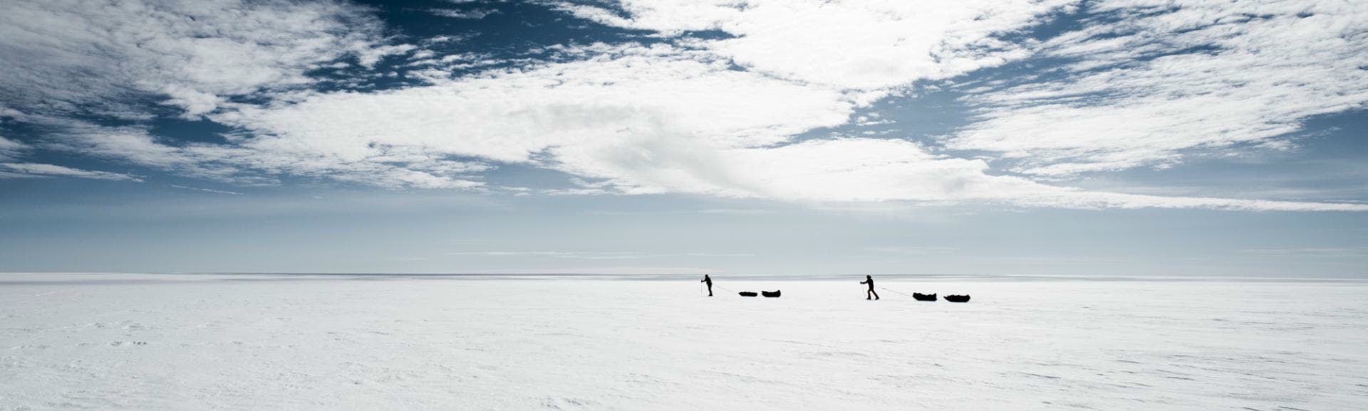 Two silhouettes pull sleds across a huge, snow-covered expanse under a dramatic sky of clouds.