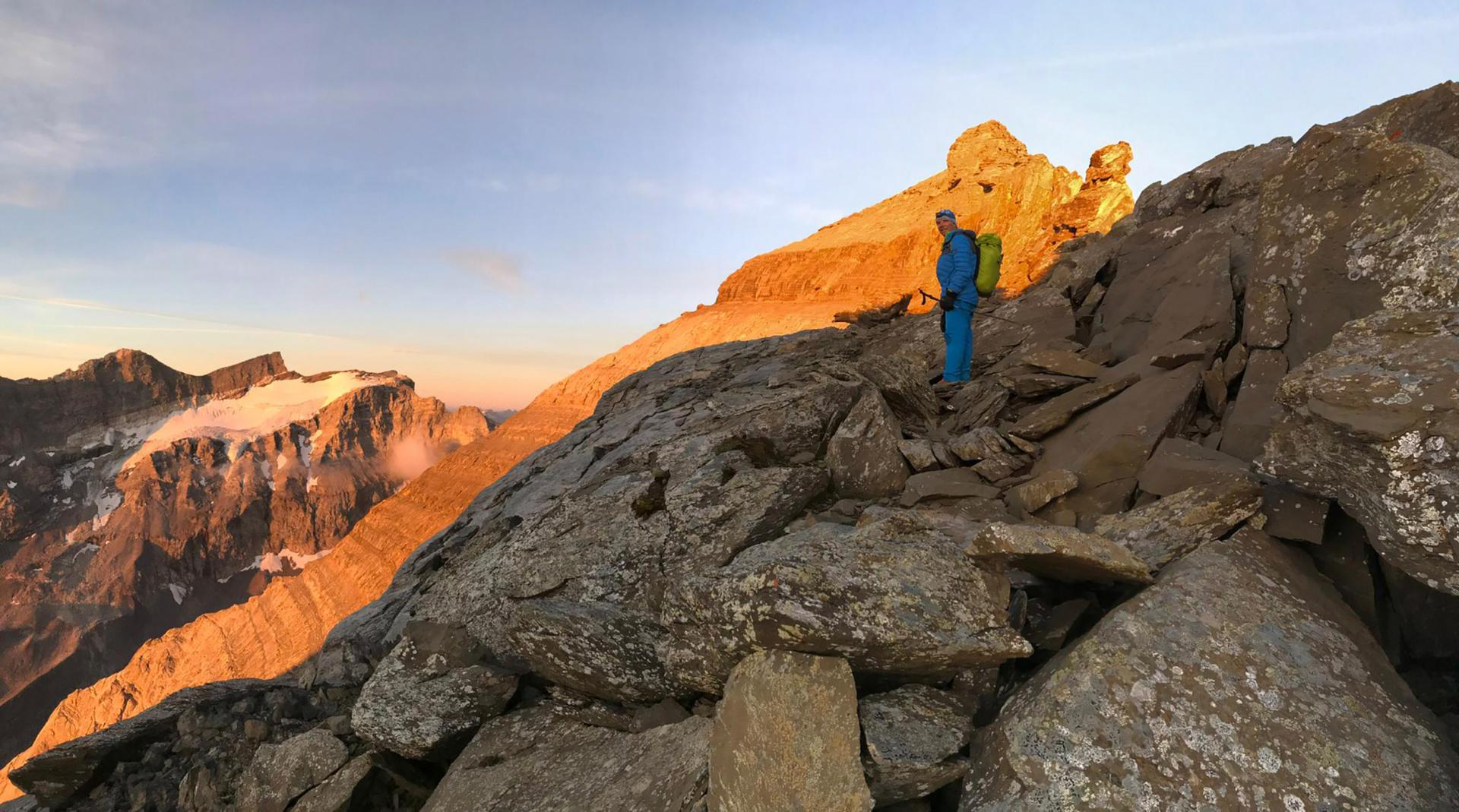 Mountaineer in blue clothes and with backpack stands on rocky ridge at sunrise, glacier in the background.