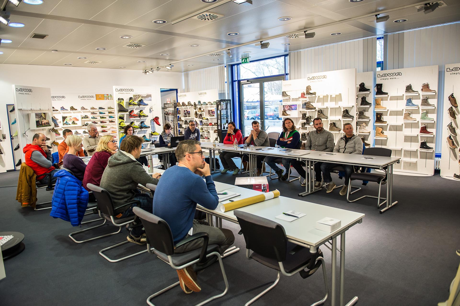 People sit at tables in a store with shoe racks in the background.