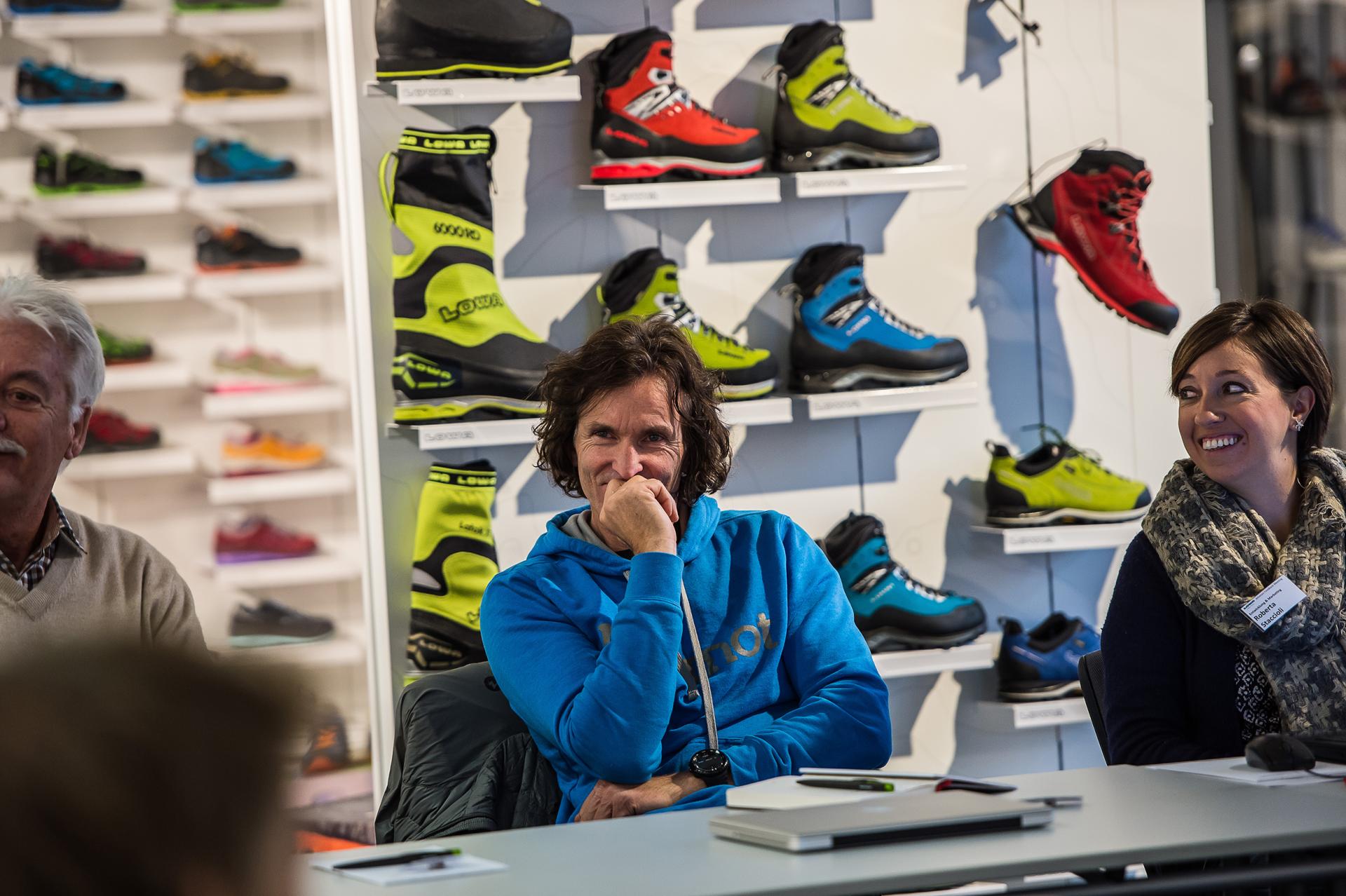 Man in blue hoodie sitting smiling next to woman with name tag in shop with many mountain boots.