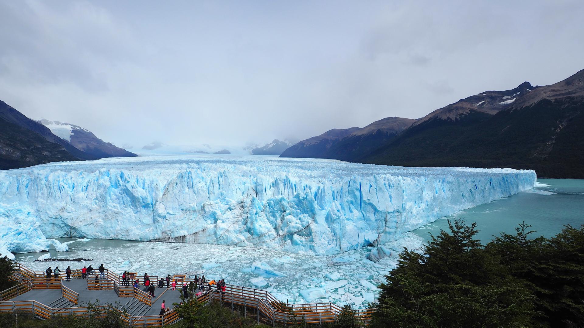Perito Moreno Glacier in Argentina with blue ice wall, floating chunks of ice and many visitors on a wooden terrace.