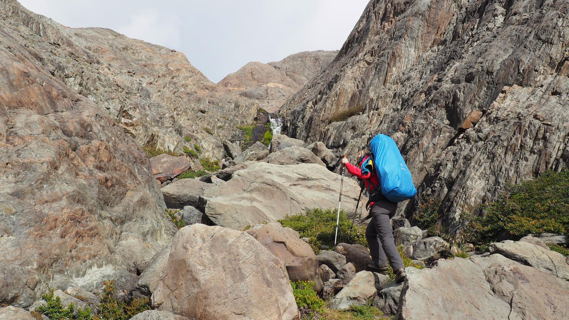 Hiker with blue backpack and poles climbing rocky slope with waterfall in background.