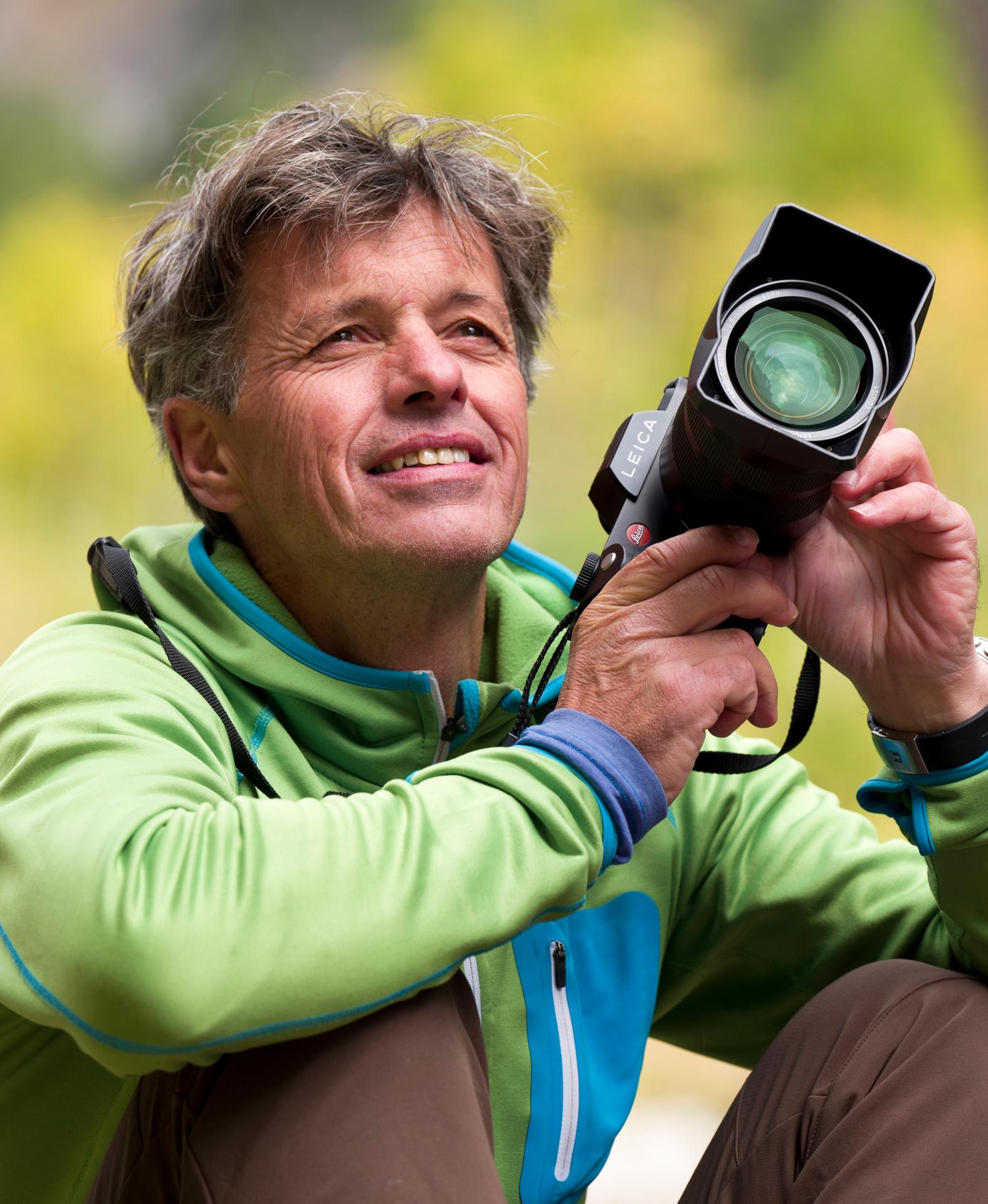 A middle-aged man with gray hair holds a Leica camera in front of his face in a green hoodie.