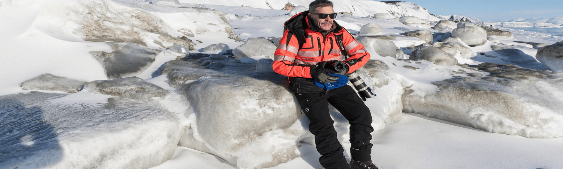 Ein Mann mit Kamera sitzt auf eisbedeckten Felsen, trägt eine orangefarbene Jacke und Sonnenbrille, umgeben von Schnee und Eis.