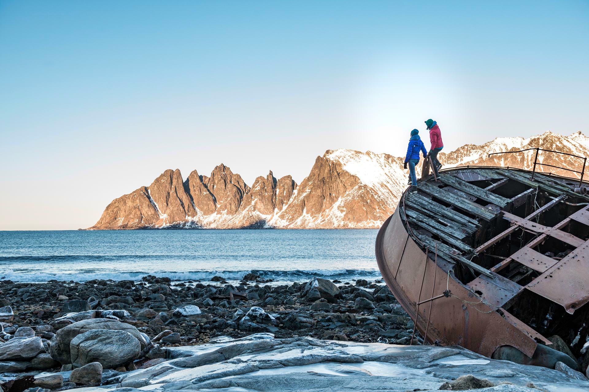Two people are standing on a shipwreck on the rocky shore, with snow-capped mountains and blue skies in the background.