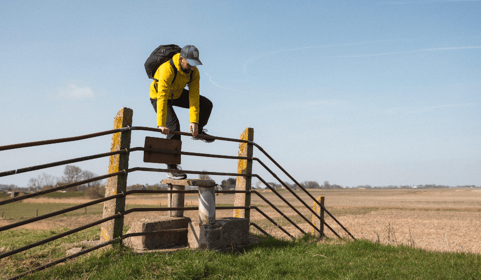 L\'uomo in giacca gialla scavalca un cancello di fronte a un ampio campo e al cielo blu.