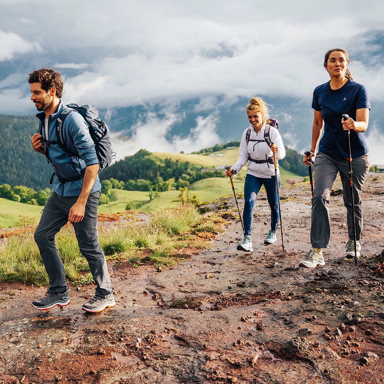 Drei Wanderer mit Rucksäcken und Wanderstöcken genießen einen Bergpfad mit Blick auf grüne Hügel und Bäume. Sie lächeln und scheinen eine gute Zeit zu haben.