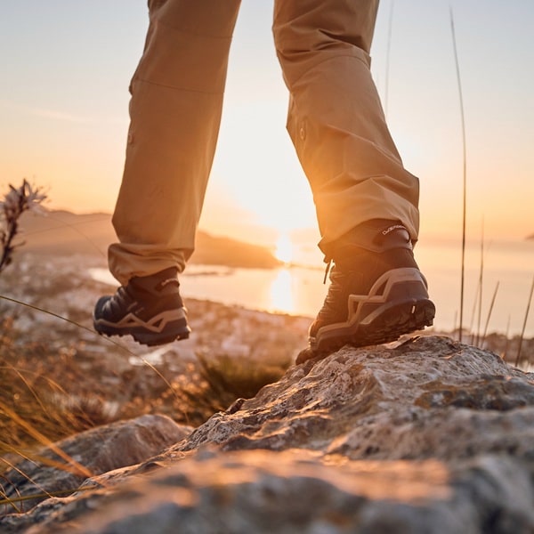 A person in hiking boots stands on a rock, the sun setting in the background.