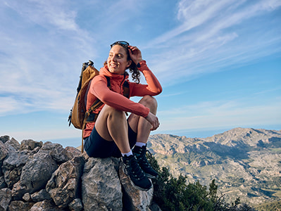 A hiker takes a break, sitting atop a rocky summit to admire a beautiful mountain range in the distance. She wears hiking boots and carries a backpack.