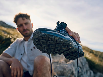 Mann hält schmutzigen Wanderschuh in die Kamera, Sonne und Berge im Hintergrund.