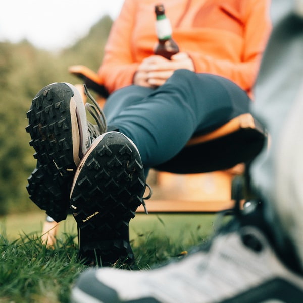 Close-up of hiking boots with rough profile, a person in an orange sweater is holding a beer bottle.