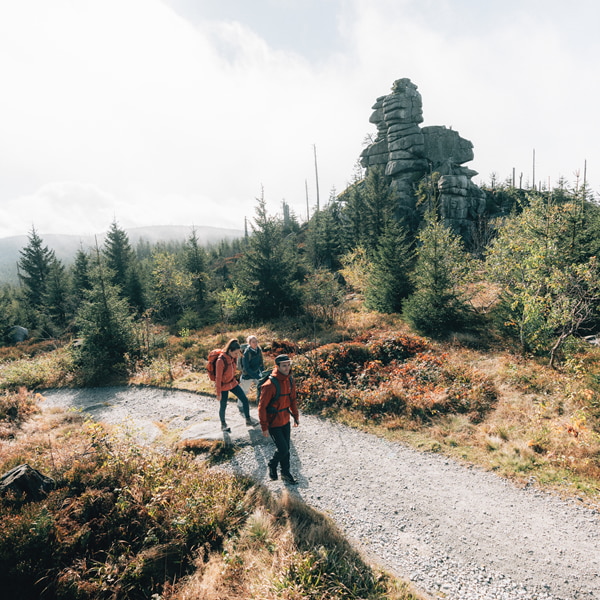 Three hikers walk on a gravel path through an autumnal mountain forest with large rock formations in the background.