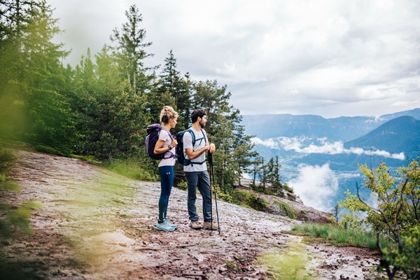 A man and a woman with backpacks look at a mountain landscape with clouds.