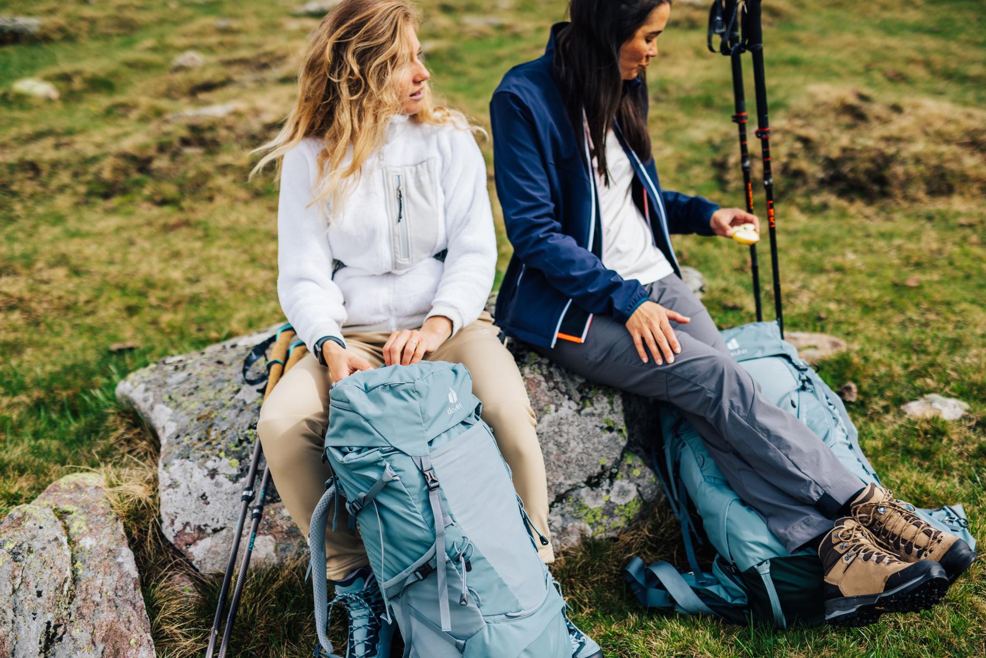 Two hikers sit on a rock with backpacks and hiking poles.