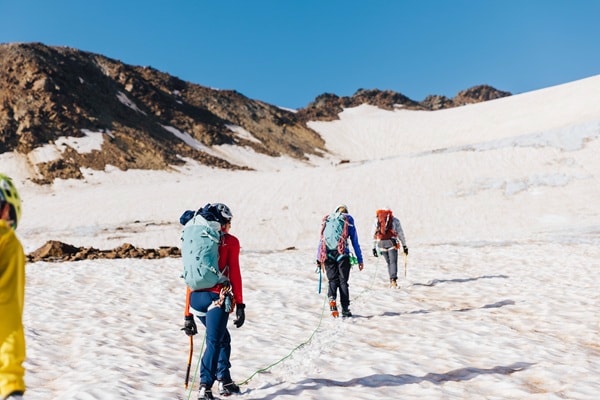 Three mountaineers with backpacks walk across a snow-covered landscape towards a mountain.