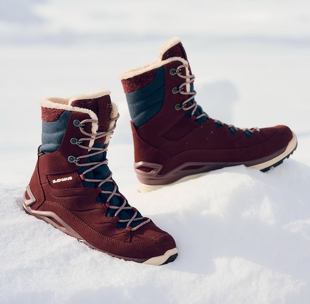 A person wearing hiking boots and black trousers is walking uphill on a rocky path, with trees and large rock formations visible in the background.