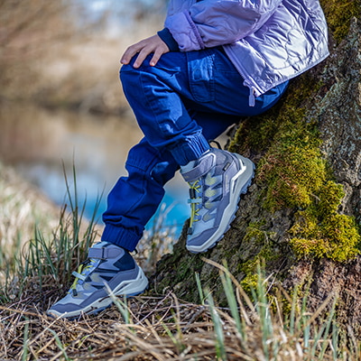 Ein Kind sitzt in blauen Hosen und Wanderschuhen aus grauem Wildleder auf einem moosbewachsenen Baumstamm.
