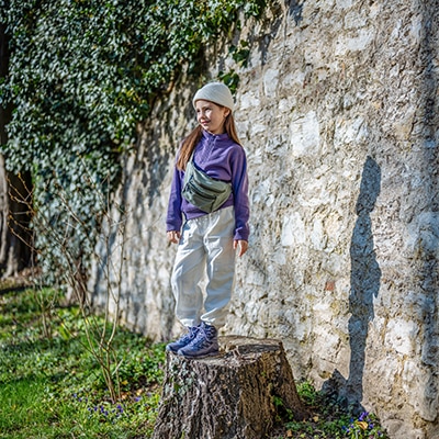 Mädchen mit Bauchttasche steht auf Baumstumpf vor Steinmauer und Bäumen.