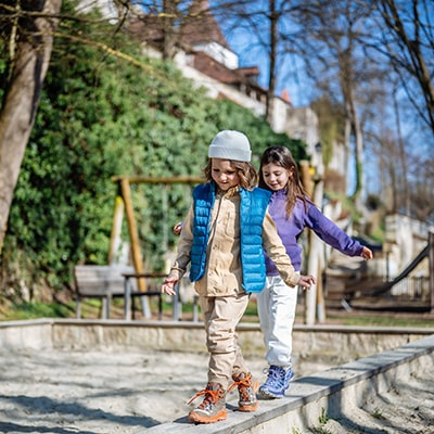 Zwei Kinder balancieren auf einem Steinweg im Freien, im Hintergrund Büsche und Gebäude.