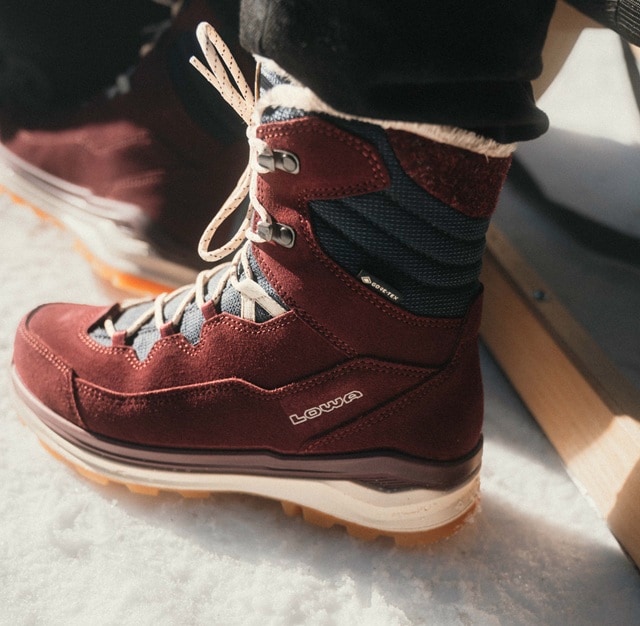 Close-up of a dark hiking boot on a rocky path, with two other people and piles of stones in the blurred background.