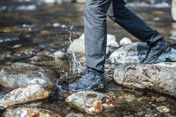 Eine Person in dunkler Hose und Wanderschuhen steigt in einen flachen, steinigen Bach und spritzt Wasser.