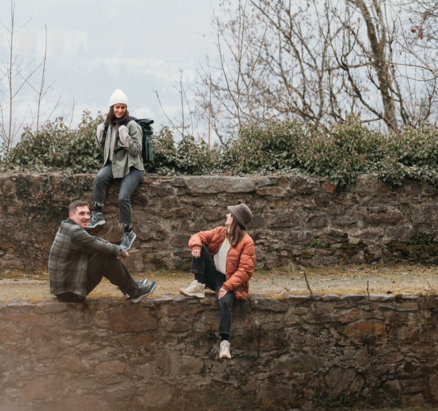Drei Personen in Outdoorkleidung entspannen sich im Freien auf einer Steinmauer.