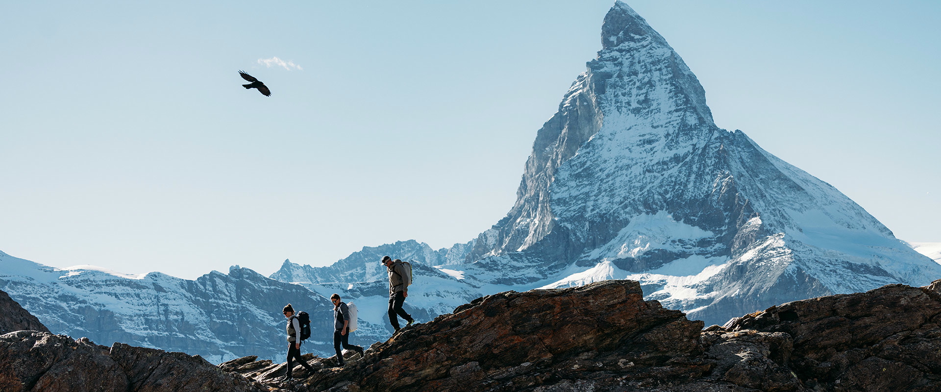 Three people are hiking on a trail through a mountainous landscape. Horses are standing beside the path.